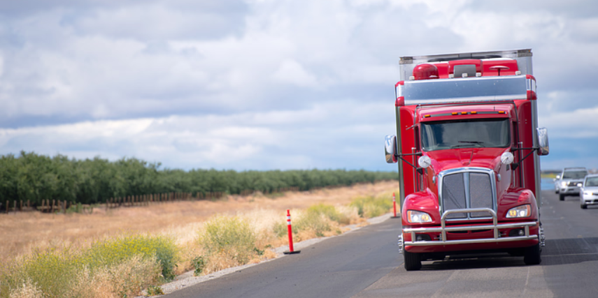 A red diesel truck drives down a country highway on a partly cloudy day.