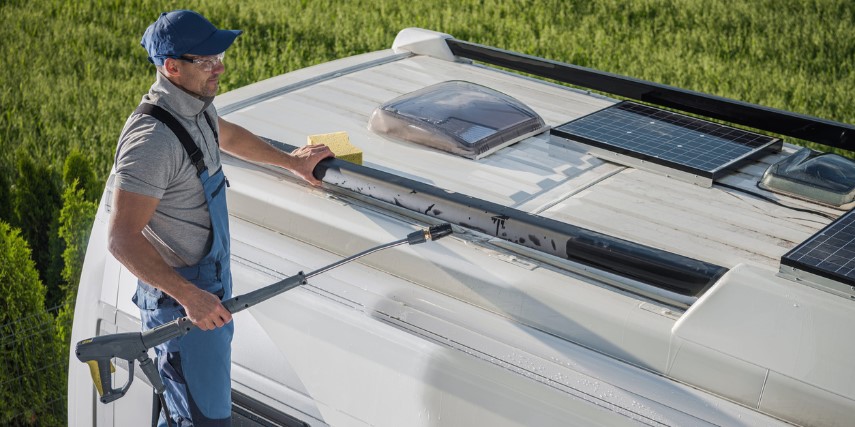 A man in a hat stands on a small ladder cleaning the top of an RV.